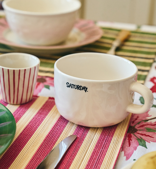 A white ceramic mug with the word 'SATURDAY.' printed in black on the side, placed on a striped pink and beige placemat.