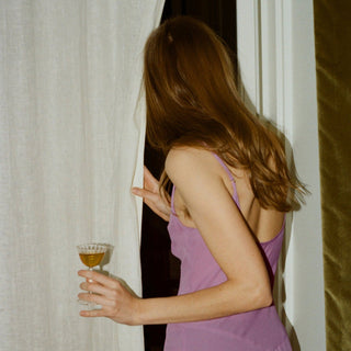 Woman in a purple dress holding a cocktail in a glass, standing behind a curtain.